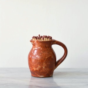 Op de afbeelding: Een kleine, rustieke, bruine keramische kruik met een handvat, gevuld met houten lucifers. De kruik heeft een gespikkelde textuur en een enigszins onregelmatige vorm, wat suggereert dat hij handgemaakt is. De lucifers hebben bruine koppen.