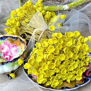 May include: A bunch of yellow flower-shaped glass beads on wire stems. The beads are arranged in a bouquet shape and are displayed on a white lace fabric background.