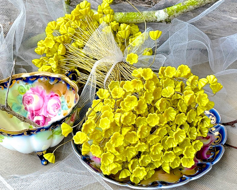 May include: A bunch of yellow flower-shaped glass beads on wire stems. The beads are arranged in a bouquet shape and are displayed on a white lace fabric background.