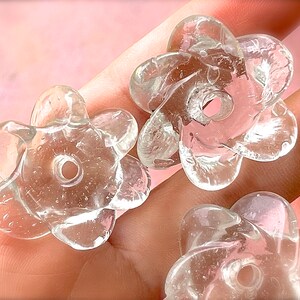 Peut inclure: Trois perles en verre transparent et incolore en forme de fleurs &agrave; cinq p&eacute;tales. Chaque perle pr&eacute;sente une surface lisse et arrondie avec un trou circulaire central pour l'enfilage. Le verre contient de fines bulles d'air internes.