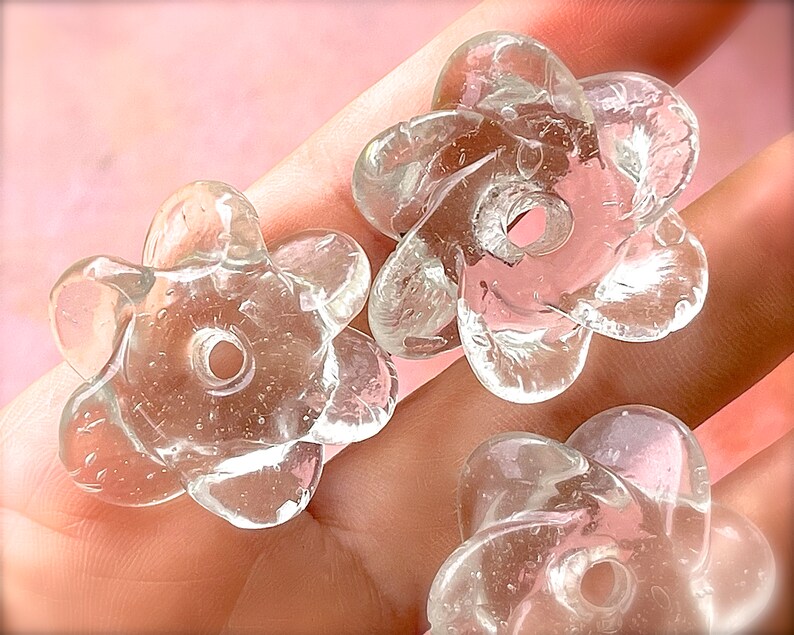 Peut inclure: Trois perles en verre transparent et incolore en forme de fleurs &agrave; cinq p&eacute;tales. Chaque perle pr&eacute;sente une surface lisse et arrondie avec un trou circulaire central pour l'enfilage. Le verre contient de fines bulles d'air internes.