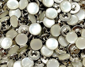 May include: A close-up of a pile of white pearl buttons with silver metal backs. The buttons are round and have a smooth, shiny finish.