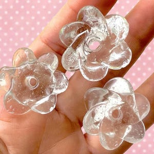 Peut inclure: Trois perles transparentes en forme de fleur avec une finition lisse et brillante. Chaque perle pr&eacute;sente cinq p&eacute;tales arrondis et un trou circulaire central pour l'enfilage. Le mat&eacute;riau semble &ecirc;tre du verre ou de l'acrylique.
