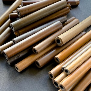 May include: A collection of cylindrical bamboo tubes in various shades of brown, from light tan to dark chocolate. The tubes have a hollow center and are arranged in a scattered pile, showcasing their natural wood grain and texture.