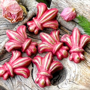 May include: Six pink and gold fleur-de-lis shaped decorative ornaments. The ornaments have a textured surface and are arranged on a wooden surface.