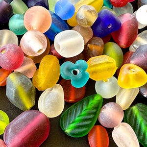 May include: A collection of colorful glass beads in various shapes and sizes. The beads are in shades of red, blue, yellow, green, and clear. Some beads are round, others are oval, and some are leaf-shaped. The beads are scattered across a dark surface.
