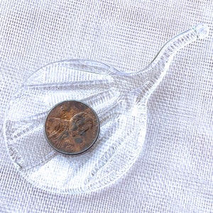 Peut inclure: Un plat en verre transparent avec une longue poign&eacute;e incurv&eacute;e. Le plat a une surface textur&eacute;e et une pi&egrave;ce de monnaie repose au centre.