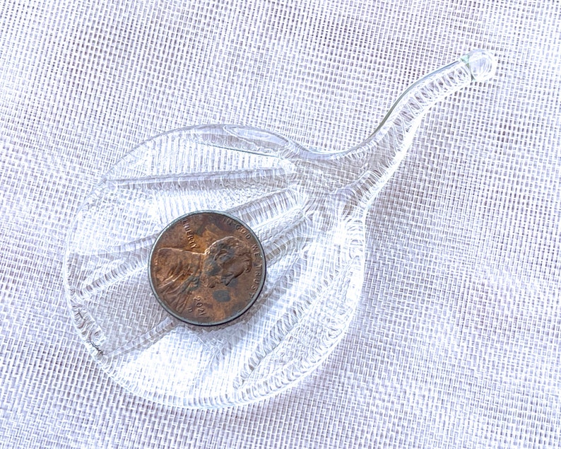 Peut inclure: Un plat en verre transparent avec une longue poign&eacute;e incurv&eacute;e. Le plat a une surface textur&eacute;e et une pi&egrave;ce de monnaie repose au centre.