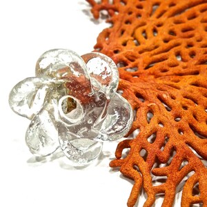 Peut inclure: Une perle en verre transparent et incolore en forme de fleur &agrave; six p&eacute;tales avec un trou central. Le verre pr&eacute;sente une surface textur&eacute;e avec des bulles et une finition brillante. La perle est plac&eacute;e &agrave; c&ocirc;t&eacute; d'un grand corail &eacute;ventail orange.