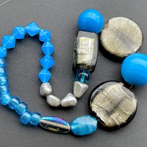 Peut inclure: Un brin de perles en verre bleu de différentes formes et tailles, y compris rondes, carrées et ovales. Les perles sont enfilées sur un fil transparent et disposées de manière aléatoire. Il y a aussi plusieurs perles plus grandes et plus décoratives dans le brin, y compris une perle en verre bleu avec un motif en spirale et une perle argentée avec une surface texturée.