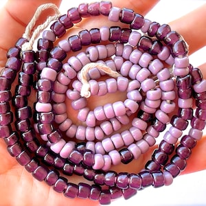 May include: A strand of purple and light purple glass beads. The beads are small and round, and they are strung on a white thread.