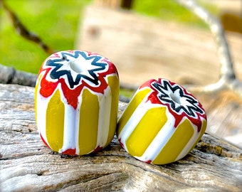 Puede incluir: Dos cuentas decorativas de vidrio. Cada cuenta es cilíndrica con un diseño de estrella en negro, rojo y blanco en la parte superior. Los lados presentan rayas verticales amarillas y blancas. Estas cuentas son probablemente para la fabricación de joyas o manualidades.