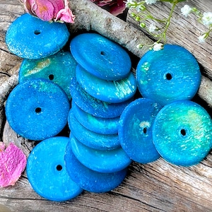 May include: A collection of round, turquoise blue wooden beads with a hole in the center. The beads have a mottled, textured surface and are arranged on a piece of weathered wood.