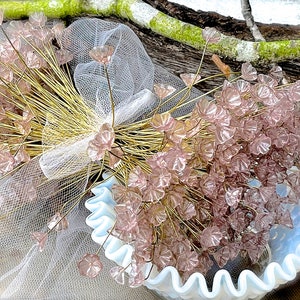 Op de afbeelding: Een boeket van delicate roze glazen bloemen met gouden stelen, gerangschikt in een witte gekrulde kom. De bloemen worden omringd door een doorschijnend wit sluier.