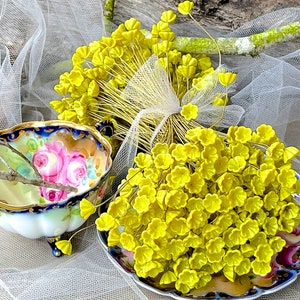May include: A bunch of yellow silk flower buds on wire stems, arranged on a blue and white porcelain plate with floral designs. The flowers are surrounded by white tulle fabric.