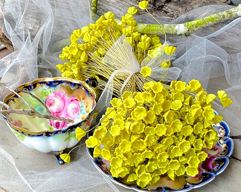 May include: A bunch of yellow silk flower buds on wire stems, arranged on a blue and white porcelain plate with floral designs. The flowers are surrounded by white tulle fabric.