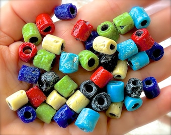 Peut inclure: Un assortiment de perles en céramique colorées dans différentes nuances de bleu, vert, rouge, jaune et noir. Les perles sont toutes de forme cylindrique et ont un trou au centre pour être enfilées.