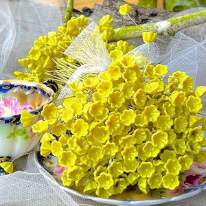 May include: A bunch of small, yellow, artificial flowers with wire stems. The flowers are arranged in a bouquet and are sitting on a white lace tablecloth.