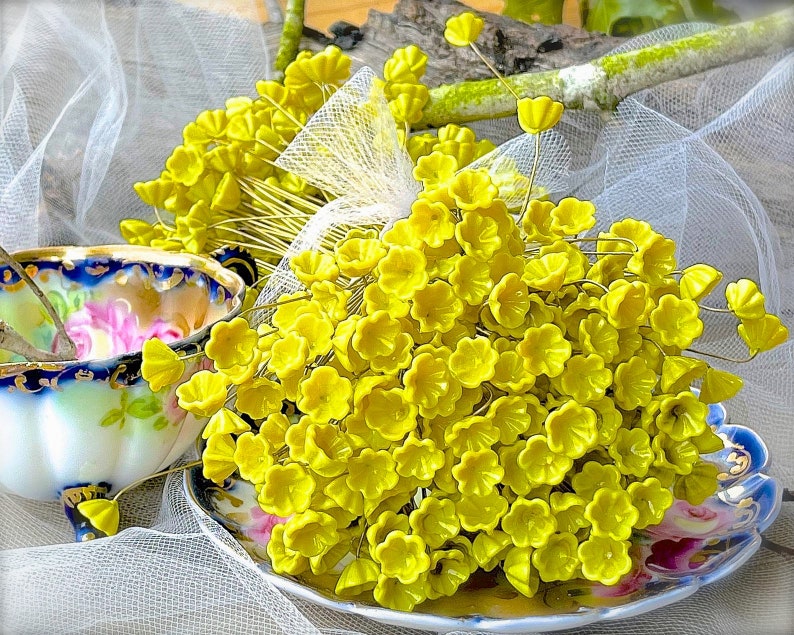 May include: A bunch of small, yellow, artificial flowers with wire stems. The flowers are arranged in a bouquet and are sitting on a white lace tablecloth.