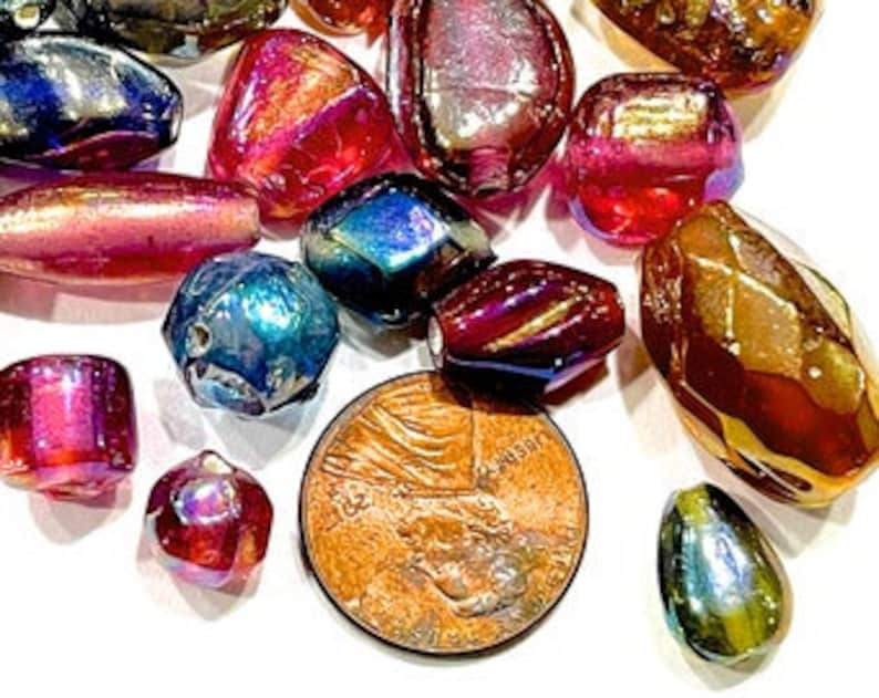 K&ouml;nnte beinhalten: Sortiment an regenbogenfarbenen Glasperlen in verschiedenen Formen und Gr&ouml;&szlig;en, darunter runde, ovale und facettierte. Die Perlen sind in Rot-, Blau-, Gr&uuml;n- und Goldt&ouml;nen gehalten. Ein Cent-St&uuml;ck ist zur Gr&ouml;&szlig;enbestimmung enthalten.
