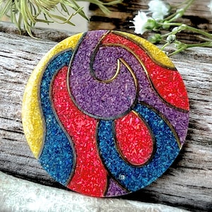May include: A round, colorful brooch with a geometric design. The brooch is made of red, blue, purple, and yellow materials with a gold outline. The brooch has a textured surface.