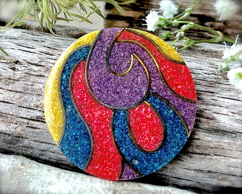 May include: A round, colorful brooch with a geometric design. The brooch is made of red, blue, purple, and yellow materials with a gold outline. The brooch has a textured surface.