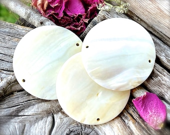 May include: Three round, off-white shell beads with two small holes each. The beads have a pearlescent sheen and are arranged on a weathered wooden surface. Dried flowers in shades of pink and white are in the background.