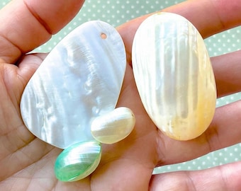 May include: Four pieces of iridescent mother-of-pearl shell components. One large irregular flat piece with a small hole, one large oval smooth piece, and two smaller rounded smooth pieces with white and light green pearlescent surfaces.