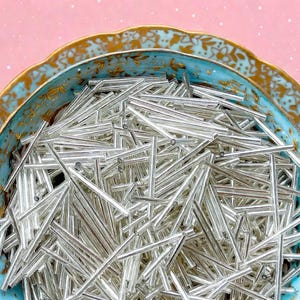 May include: A large collection of metallic silver bugle beads, each featuring a long, thin, cylindrical shape with a smooth, reflective surface, contained within a decorative bowl with a blue and gold floral pattern on the interior rim.