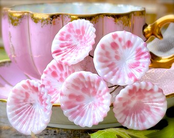 May include: Close-up of six white and pink glass flower-shaped beads. The beads have a scalloped edge and a radial design of pink spots and lines. A pink teacup with gold trim is in the background.