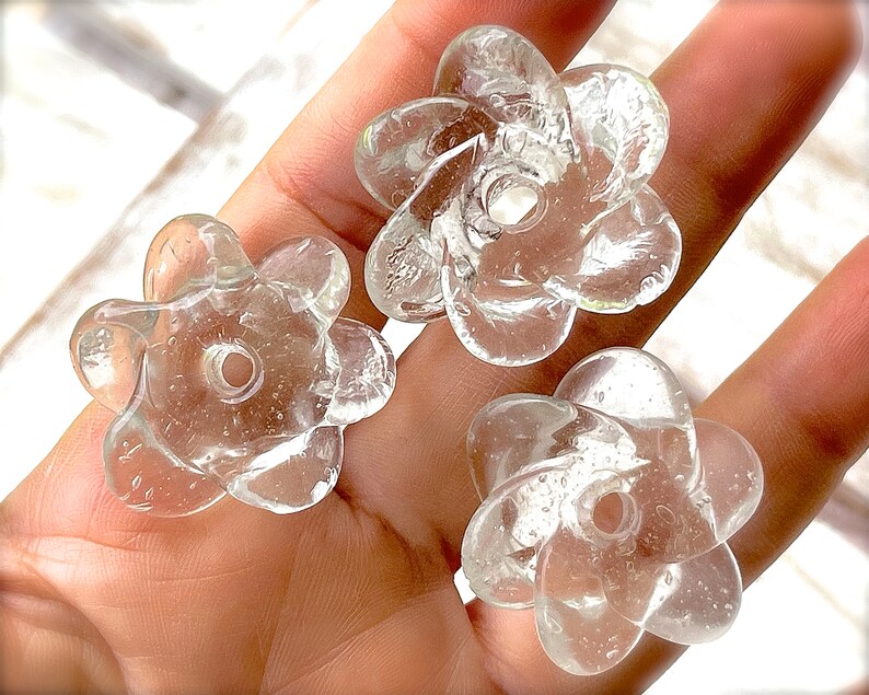 Peut inclure: Trois perles en verre transparent en forme de fleurs &agrave; cinq p&eacute;tales. Chaque perle pr&eacute;sente une surface lisse et arrondie avec de petites bulles d'air internes et un trou central circulaire pour l'enfilage, offrant un aspect transparent.