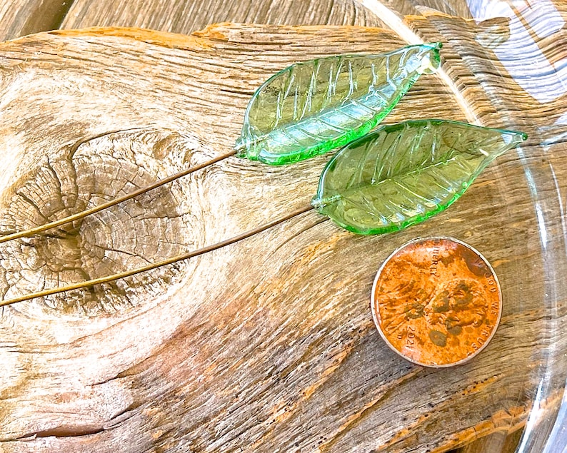 May include: Two green glass leaves on wire stems. The leaves are on a wooden surface with a copper penny.