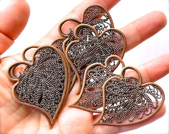 Può includere: Quattro ciondoli in metallo a forma di cuore decorati con un motivo a filigrana. I ciondoli sono di colore bronzo scuro e presentano un motivo delicato e intricato.