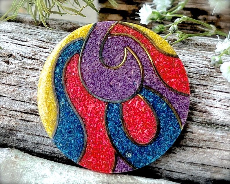 May include: A round, colorful, decorative object with a swirling pattern in shades of red, yellow, blue, and purple. The object is made of a textured material that resembles small, colorful pebbles.