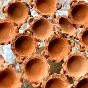 Pode incluir: Uma coleção de pequenos vasos de terracota com bordas festonadas e duas alças. Os vasos estão dispostos em um padrão circular sobre uma superfície de cor clara.
