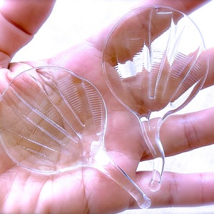 Peut inclure: Deux cuill&egrave;res en plastique transparentes avec un motif en &eacute;ventail. Les cuill&egrave;res sont tenues dans une main.