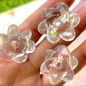 Peut inclure: Trois perles en verre transparent en forme de fleurs &agrave; cinq p&eacute;tales. Chaque perle pr&eacute;sente une surface lisse et translucide avec un trou circulaire central pour l'enfilage. Le verre est incolore et refl&egrave;te la lumi&egrave;re avec de fines bulles.
