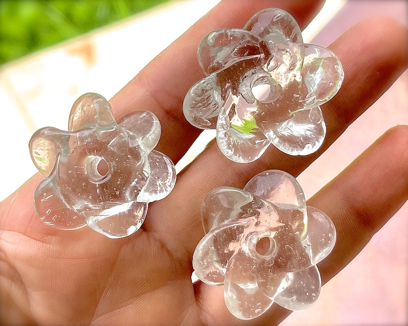 Peut inclure: Trois perles en verre transparent en forme de fleurs &agrave; cinq p&eacute;tales. Chaque perle pr&eacute;sente une surface lisse et translucide avec un trou circulaire central pour l'enfilage. Le verre est incolore et refl&egrave;te la lumi&egrave;re avec de fines bulles.