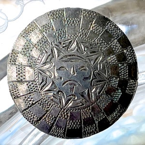 May include: A silver circular button with a stylized face in the center surrounded by a geometric pattern.