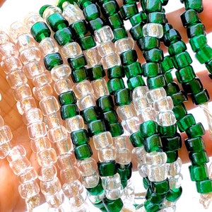May include: Strands of clear and emerald green glass beads. The beads are small, square-shaped, and strung on a natural-colored cord. The clear beads alternate with the green beads, creating a striped pattern. The beads are held up against a white background.