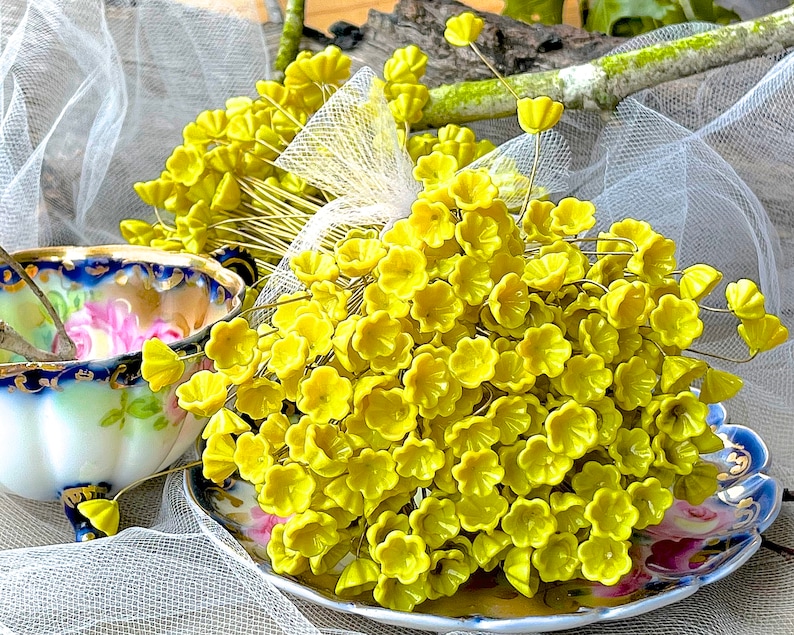 May include: A bunch of small, bright yellow flower-shaped embellishments on wire stems. The embellishments are clustered together and arranged on a white lace fabric background.