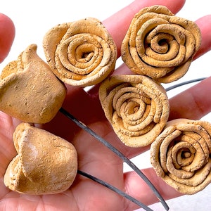 Puede incluir: Seis pequeñas rosas de cerámica beige con tallos de alambre. Las rosas están dispuestas en una mano, con los tallos extendiéndose hacia afuera.