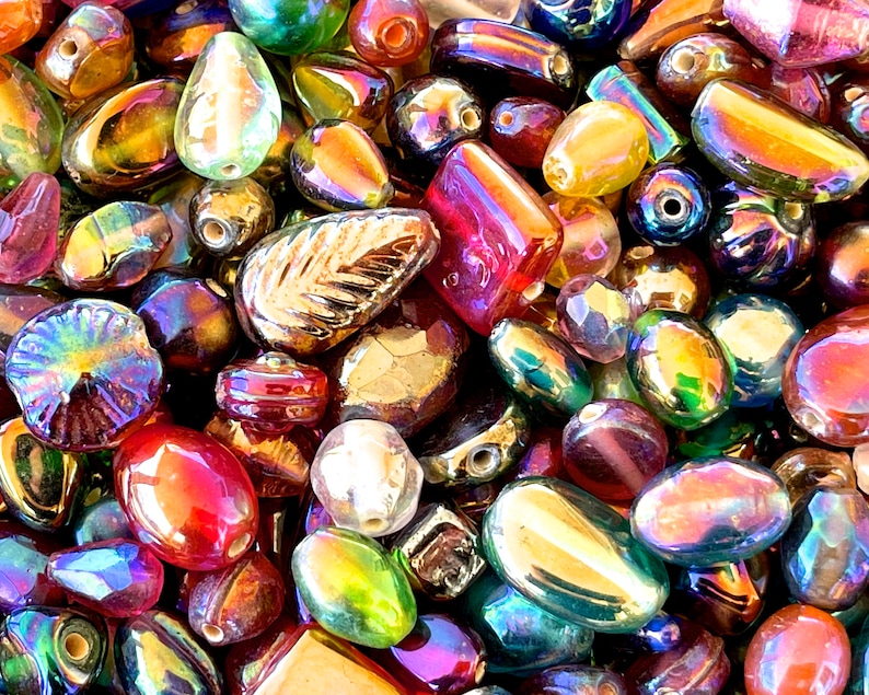 K&ouml;nnte beinhalten: Eine Sammlung von regenbogenfarbenen Glasperlen in verschiedenen Formen und Gr&ouml;&szlig;en, darunter runde, ovale und blattf&ouml;rmige Perlen. Die Perlen haben einen regenbogenartigen Glanz und sind perfekt f&uuml;r die Schmuckherstellung oder das Basteln.