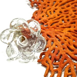 Peut inclure: Un objet en verre souffl&eacute; transparent en forme de fleur &agrave; plusieurs p&eacute;tales tourbillonnants avec une ouverture circulaire centrale, pr&eacute;sentant de petites bulles d'air internes dans tout le verre, plac&eacute; &agrave; c&ocirc;t&eacute; d'un grand corail &eacute;ventail orange.
