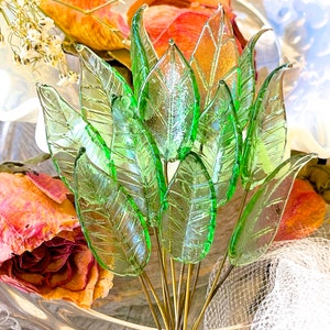 May include: A bouquet of green glass leaves with a delicate, translucent texture. The leaves are arranged in a fan shape and are held together by a wire stem. The leaves are surrounded by dried flowers and a white lace fabric.