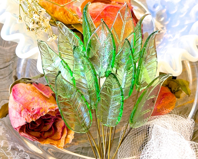 May include: A bouquet of green glass leaves with a delicate, translucent texture. The leaves are arranged in a fan shape and are held together by a wire stem. The leaves are surrounded by dried flowers and a white lace fabric.