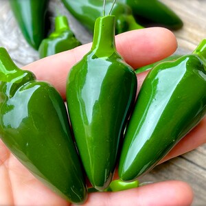May include: Four green ceramic pepper ornaments with a shiny finish. Each pepper has a silver wire loop for hanging.