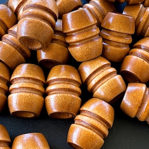 May include: A collection of small, barrel-shaped wooden beads. The beads are a warm, medium brown color with a glossy finish. Each bead has horizontal grooves carved around its circumference, adding texture and detail. The beads are scattered, creating a sense of abundance.