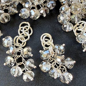 Puede incluir: Primer plano de pendientes plateados con múltiples cuentas de cristal transparente facetadas. Los pendientes tienen un diseño de racimo, con cada cuenta conectada por pequeños eslabones plateados. Los cristales reflejan la luz, creando un efecto brillante.