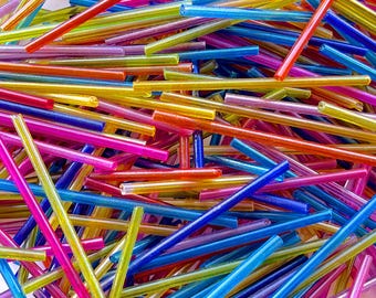 May include: A close-up shot of a pile of colorful, thin, glass rods. The rods are in various colors, including red, orange, yellow, green, blue, and pink. The rods are scattered and overlapping, creating a vibrant and textured surface.